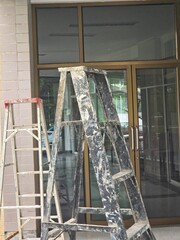 A construction or renovation scene showing two ladders, one black and one white, positioned in front of a glass door entrance.