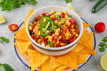 Corn salsa in a bowl with lime, jalapeno pepper, fresh herbs and tortilla chips on a concrete background. Mexican cuisine. Healthy food.