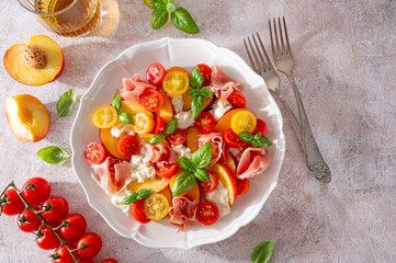 Salad with mozzarella, peaches, prosciutto ham, tomatoes and fresh basil on a white plate on a concrete background. Top view, copy space. Hard light.