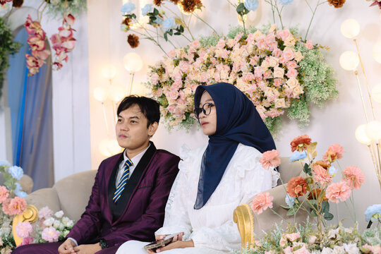 An Asian man and a hijab-wearing woman with glasses sit together on a sofa wearing elegant formal suits, with a beautiful floral backdrop behind them. Their poised expressions and stylish appearance