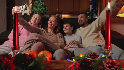 Happy family in pajamas making video call on Christmas Eve sitting on sofa in cozy room with Christmas tree in background, red candles and tangerines on table. - Powered by Adobe