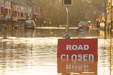 A 'Road Closed' sign partially covered in flood water lit by the evening sun