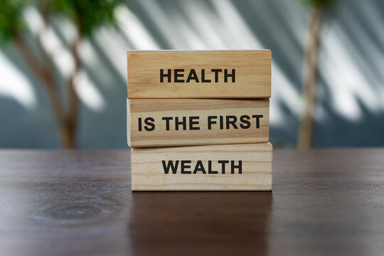 Wooden blocks stacked with inspirational quote "Health is the first wealth" on a wooden table, with blurred natural background and sunlight.
