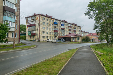 street in Korsakov, Sakhalin Island, Russia, lined with Soviet-era apartment blocks, green trees, and parked cars on a cloudy day.
