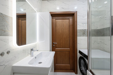 A bright bathroom features a white sink with a chrome faucet, a lit mirror, and a wood door. Marble-like tiles adorn the walls, with a glimpse of a washing machine behind a shower enclosure
