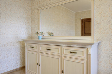 A cream-colored dresser with a large mirror sits against a patterned wall. A small blue vase with flowers is on top