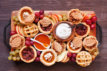 Autumn pie theme dessert board. Top view against a dark wood background. Pumpkin, apple and pecan mini pies, with whipped cream, caramel and fruit.