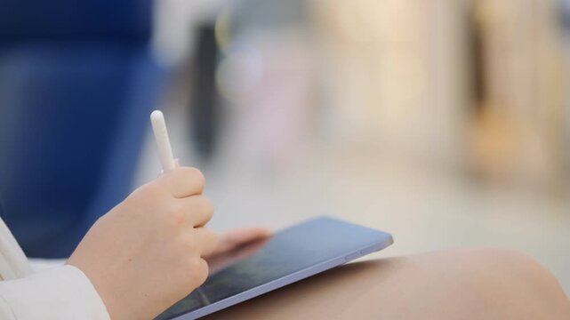 Person is writing on a tablet with a pen. The tablet is on a table. The person is sitting on a chair