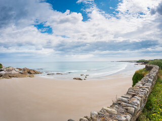 Wide view of Playa de las Catedrales in Galicia, Spain, showing sandy beach, rocky formations, calm waves, stone wall, and dramatic cloudy sky along the Atlantic coast.