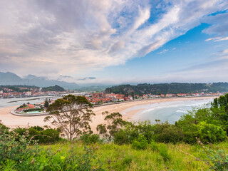 Coastal town of Ribadesella in Asturias, Spain, with sandy river mouth curving into the sea, colorful houses, mountains in background, cloudy sky above, blending culture and nature.