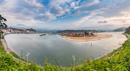 Coastal town of Ribadesella in Asturias, Spain, with sandy river mouth curving into the sea, colorful houses, mountains in background, cloudy sky above, blending culture and nature.