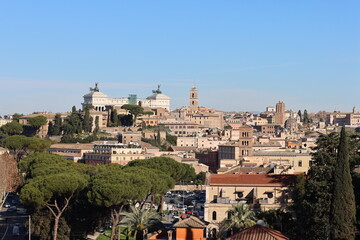Fototapeta premium Panoramic view of Rome with historic buildings, domes, towers, and the Vittoriano monument under a clear blue sky, framed by green trees.