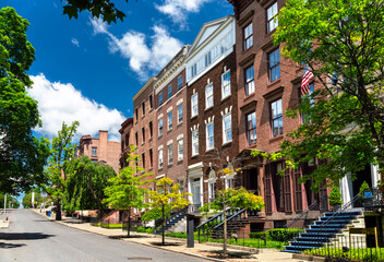 A scenic street lined with historic brick rowhouses and green trees under a bright blue sky on a sunny summer day in Albany, New York, United States