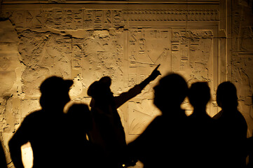 Tourists marvel at ancient Egyptian hieroglyphs illuminated by warm light on a stone wall with a guide pointing towards intricate carvings depicting historical narratives and symbols of a bygone civil