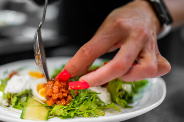 Close-up of a hand plating gourmet salad with boiled eggs, cucumber, greens, and creamy topping. Food styling, red nails, and wristwatch visible. Culinary preparation concept.