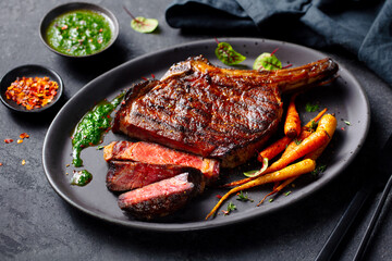 Tomahawk steak with chimichurri sauce on a black plate. Grey background. Close up.