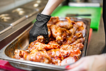 Chef hand in black glove handling marinated chicken drumsticks on metal tray in kitchen. Raw meat preparation with spices. Culinary scene with cutting board and knife in background.