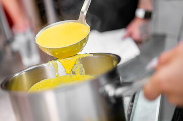 Close-up of a person ladling thick yellow soup from a stainless steel pot in a kitchen setting, highlighting texture, cooking process, and food preparation