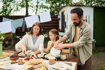 Caucasian middle aged woman slicing pie while Caucasian middle aged man pouring tea and Caucasian child girl sitting at outdoor table