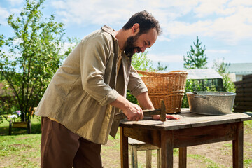 Middle aged Caucasian man working outdoors hammering nail into wooden table, standing beside wicker basket and metal basin in garden with trees and greenhouse in background