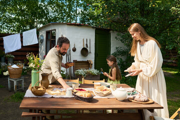 Caucasian middle aged man arranging food on outdoor table while woman preparing plates and girl using smartphone in backyard garden setting
