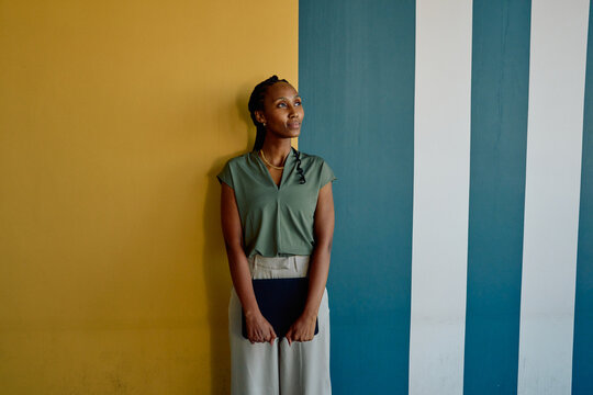African young businesswoman with tablet looking up while standing against colorful wall in office