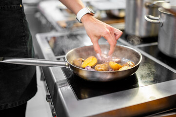 Chef sautéing halved potatoes in a steaming pan on electric stove. Professional kitchen setting, black apron, white watch, cooking at high heat