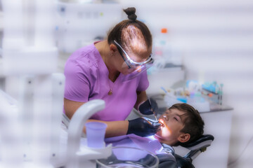 A dentist in purple scrubs examines a young boys teeth while he sits in the dental chair, wearing protective glasses and gloves during the procedure.
