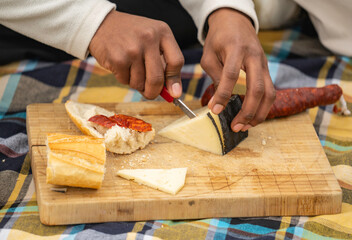 Close-up of hands cutting cheese on a wooden board, next to chorizo and bread, during a picnic