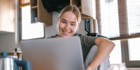 Gen Z woman working from home at cozy kitchen table with laptop and phone. Digital nomad...