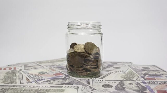 Timelapse of shiny gold coins falling into a clear glass jar on a white background, with scattered dollar bills around.