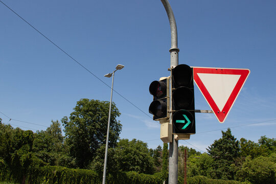 Traffic signal displaying green light for right turn, with yield sign visible, under clear blue sky, surrounded by lush greenery and urban infrastructure elements