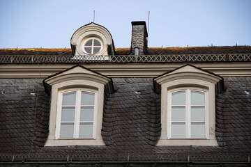 Architectural details of a historic building featuring two large windows and a small round window, showcasing intricate roof design and elegant craftsmanship in a serene sky backdrop