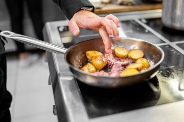 Chef sautéing octopus and halved potatoes in a stainless steel pan on a professional stovetop. Close-up of hands-on cooking in a restaurant kitchen.