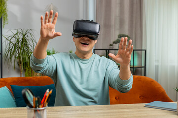 Asian man using virtual reality futuristic technology VR app headset helmet typing on virtual keyboard, play simulation 3D 360 online video game at home apartment. Guy in goggles sitting on sofa
