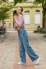 Obraz premium Happy Caucasian young woman enjoying morning coffee hot drink and smiling outdoors. Relaxing, taking a break. Girl walking through urban city center street, drinking coffee to go. Town lifestyles.
