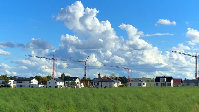 Construction cranes work to build new homes in a suburban location surrounded by vibrant greenery and scattered clouds
