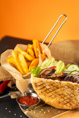 Grilled pita sandwich with fries and ketchup on wooden board, vibrant orange background
