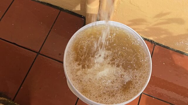 Polluted water flows freely from a tap. The water fills a white bucket standing under it. The water is brown and discolored. 
