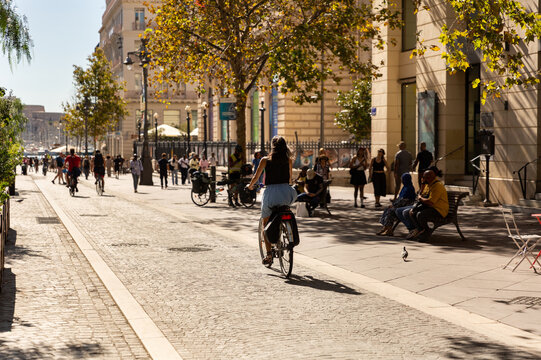 V&eacute;lo en ville, jeune femme &agrave; v&eacute;lo au premier plan vue de dos sur une piste cyclable du centre ville de Marseille, promeneurs et all&eacute;e d&rsquo;arbres au second plan