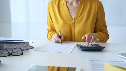 Female accountant wearing yellow shirt is sitting at white desk and using calculator while taking...