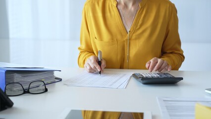 Female accountant wearing yellow shirt is sitting at white desk and using calculator while taking...