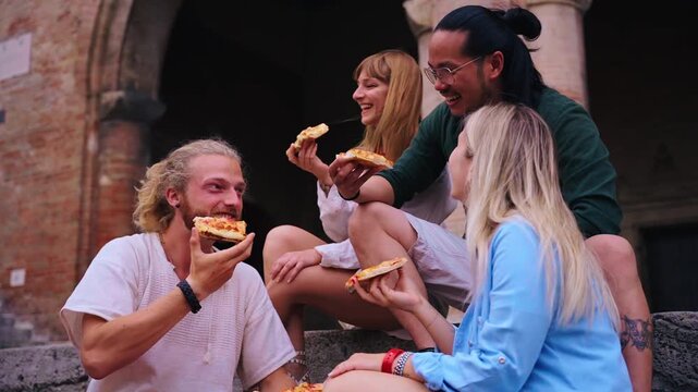 Group of cheerful young multiethnic friends are laughing and enjoying eating street food pizza together while sitting on steps in front of a historical building