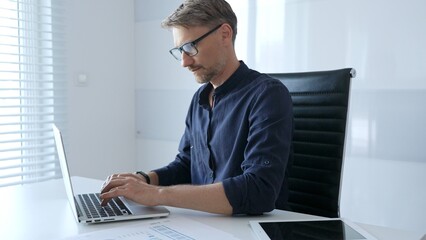 Professional businessman wearing glasses, intensely working on laptop, typing amid modern office...