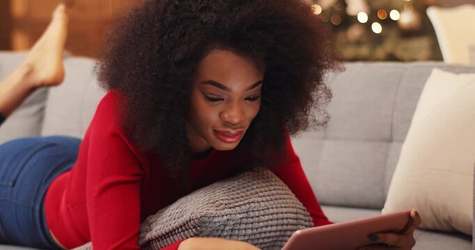 Awesome African girl in red sweater using tablet on background of fascinated Christmas tree.Young woman browsing and listening music. Indoors.