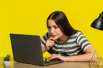 Woman sitting at table attentively reading complex material on laptop with focused expression during online learning. Girl on yellow background shows concentration engagement in serious study process