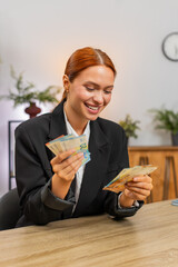 Young businesswoman at home office fans euros above desk smiling at booming earnings and freedom. Freelancer girl at table counts cash celebrating big raise bonus financial triumph investment plan