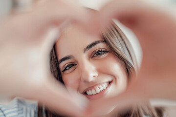 Obraz premium Beautiful woman showing heart sign with folded fingers smiling looking at camera, head shot portrait. Attractive optimistic young female demonstrate symbol of love feels happy posing alone at home