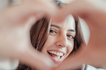 Fototapeta premium Beautiful woman showing heart sign with folded fingers smiling looking at camera, head shot portrait. Attractive optimistic young female demonstrate symbol of love feels happy posing alone at home