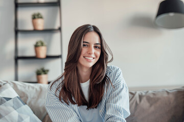 Young attractive cheerful lady sitting on sofa, relaxing looking at the camera at home. Beautiful brunette woman on her 30s chatting on video call online